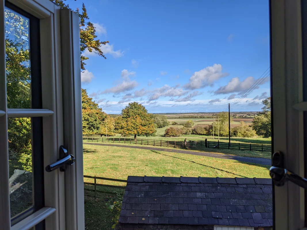 Open cottage window revealing sunlit fields, trees, and clear blue sky in rural landscape