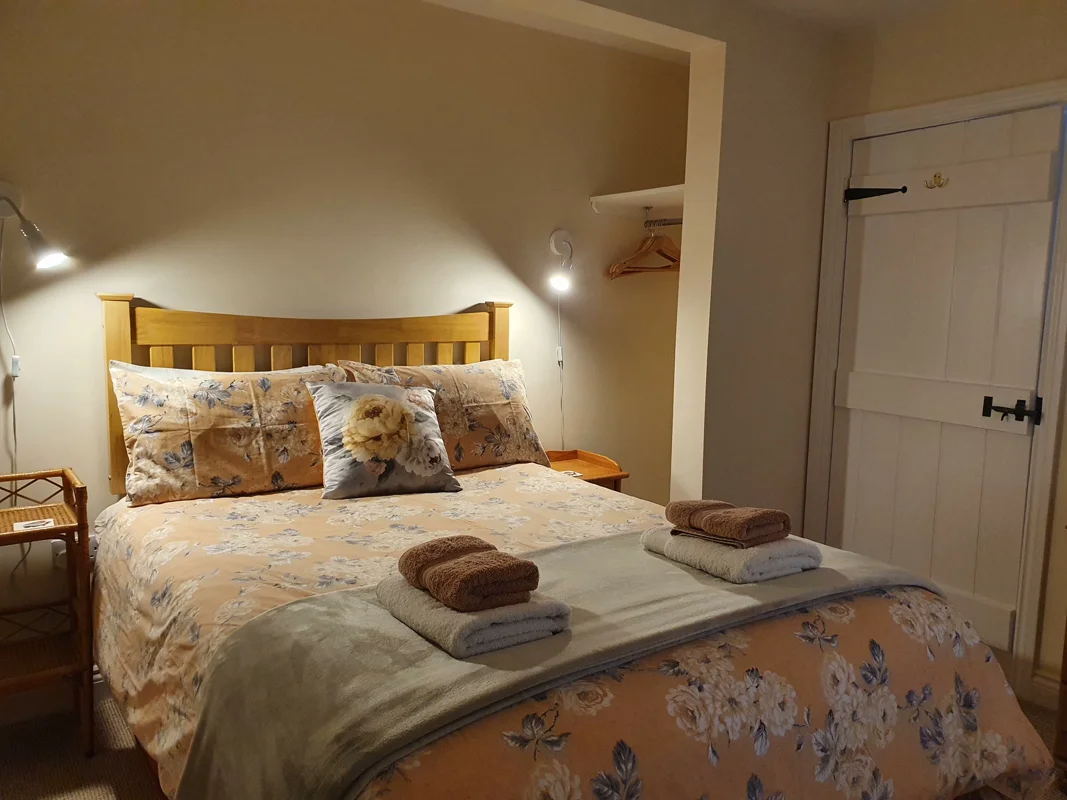 Warmly lit cottage bedroom with wooden headboard, floral bedspread, two nightstands, and stacked towels.