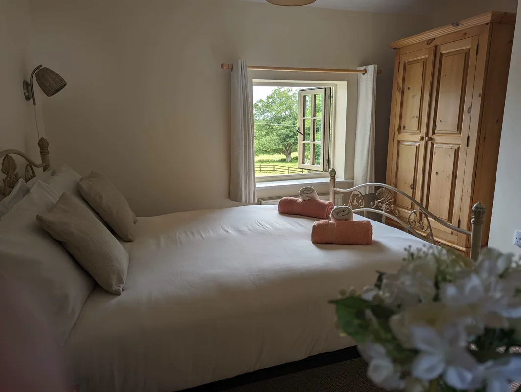 Comfortable bed, pink rolled towels, garden view through window, wooden wardrobe, bedside lamp, and floral arrangement in cottage bedroom.