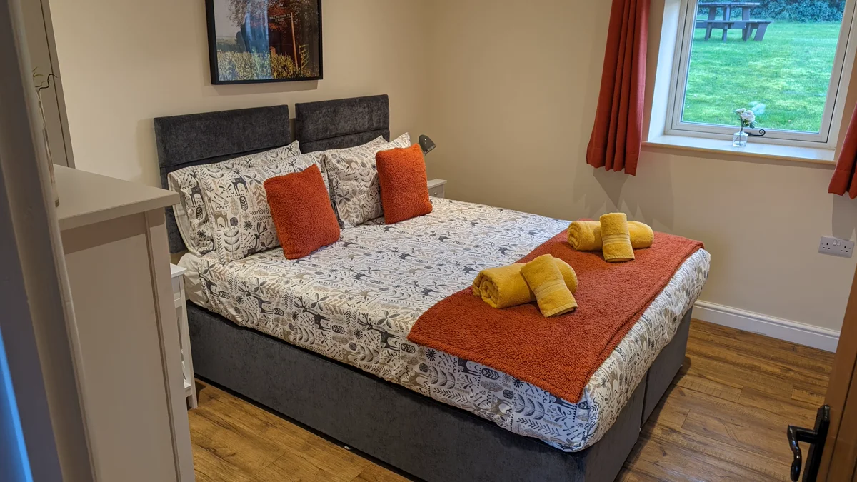 Comfortable bed with patterned duvet, orange cushions, rolled towels, and garden view through window.