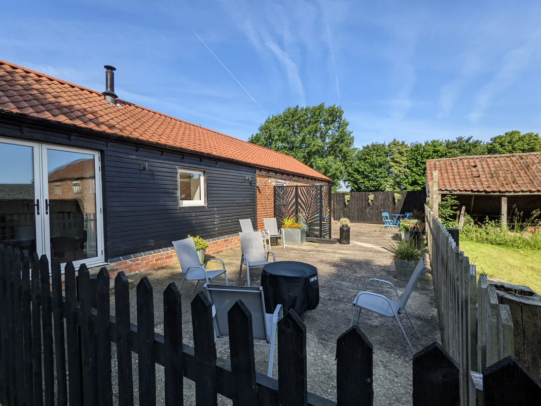 Black timber cottage with red tile roof, gravel patio, garden chairs, and lush greenery. Bright sunny day.