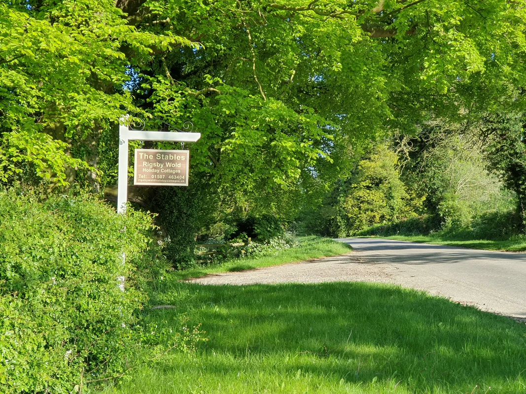 Sign for The Stables Rigsby Wald holiday cottages on tree-lined country road, lush greenery.